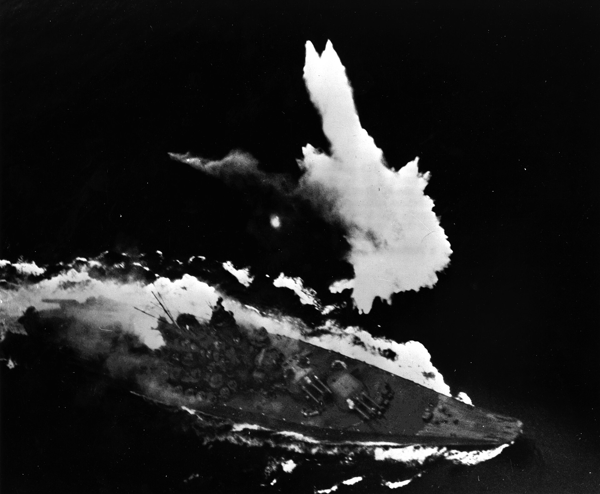 Black and white historical photo of the Yamato maneuvering under heavy aerial bombardment, surrounded by water plumes.